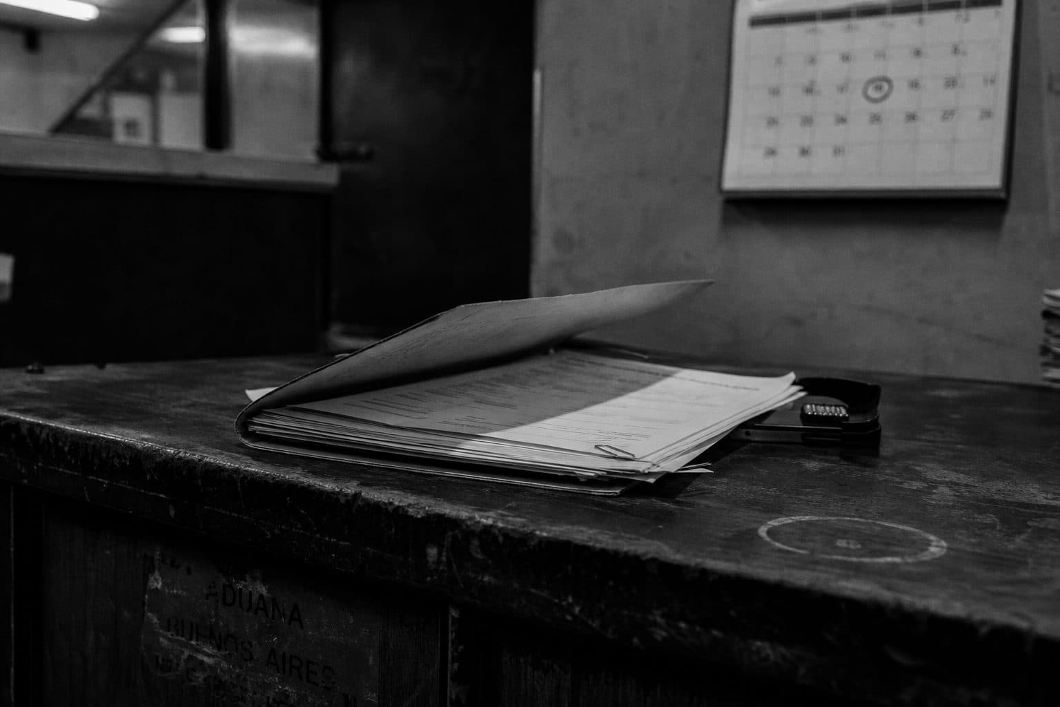 Carpeta con pólizas y formularios de la Aduana sobre un mostrador, con un calendario de pared al fondo, fotografía documental en blanco y negro.